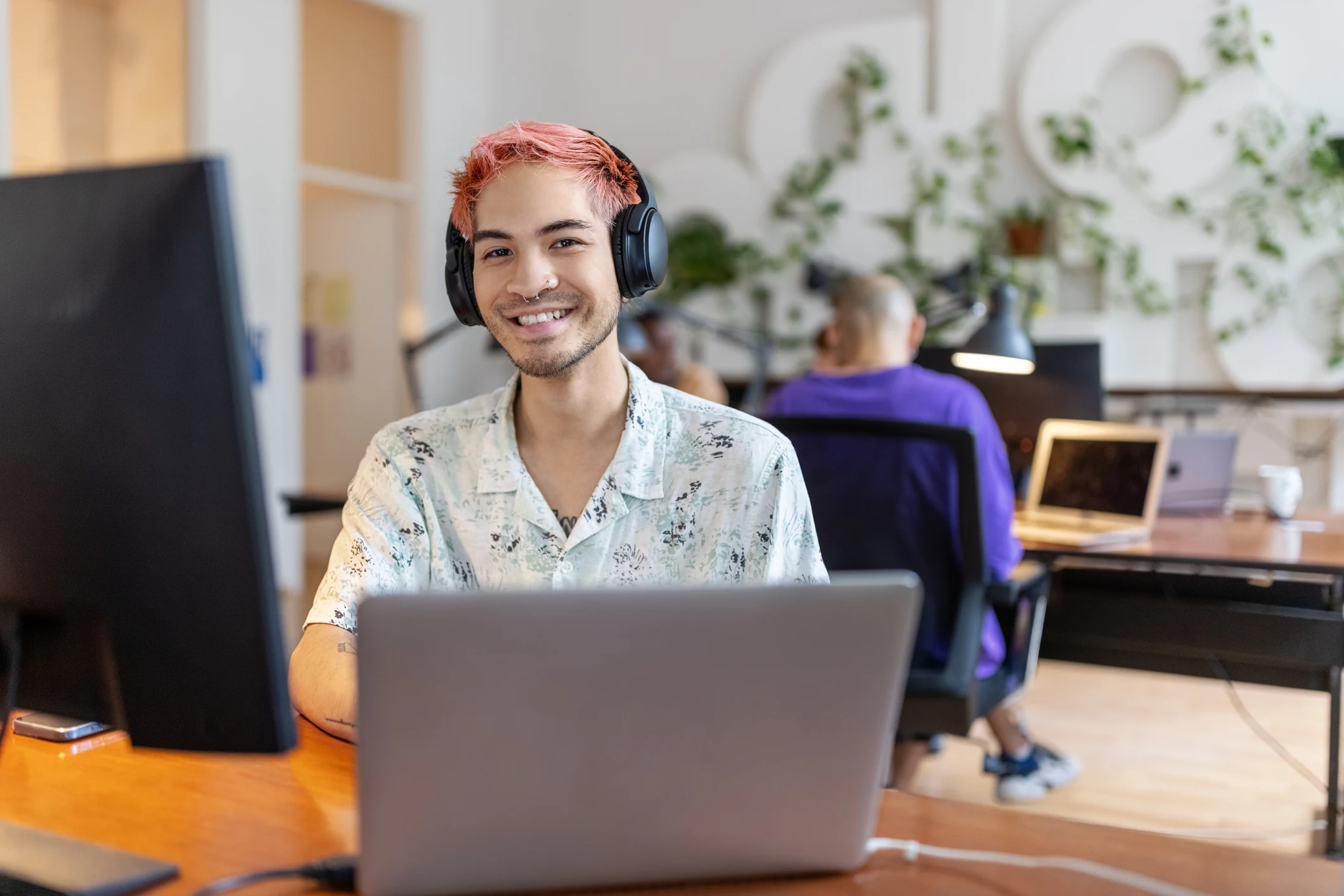 Portrait of Asian man with headphones working at startup office. Young man with dyed red hair and nose piercing looking at camera and smiling.