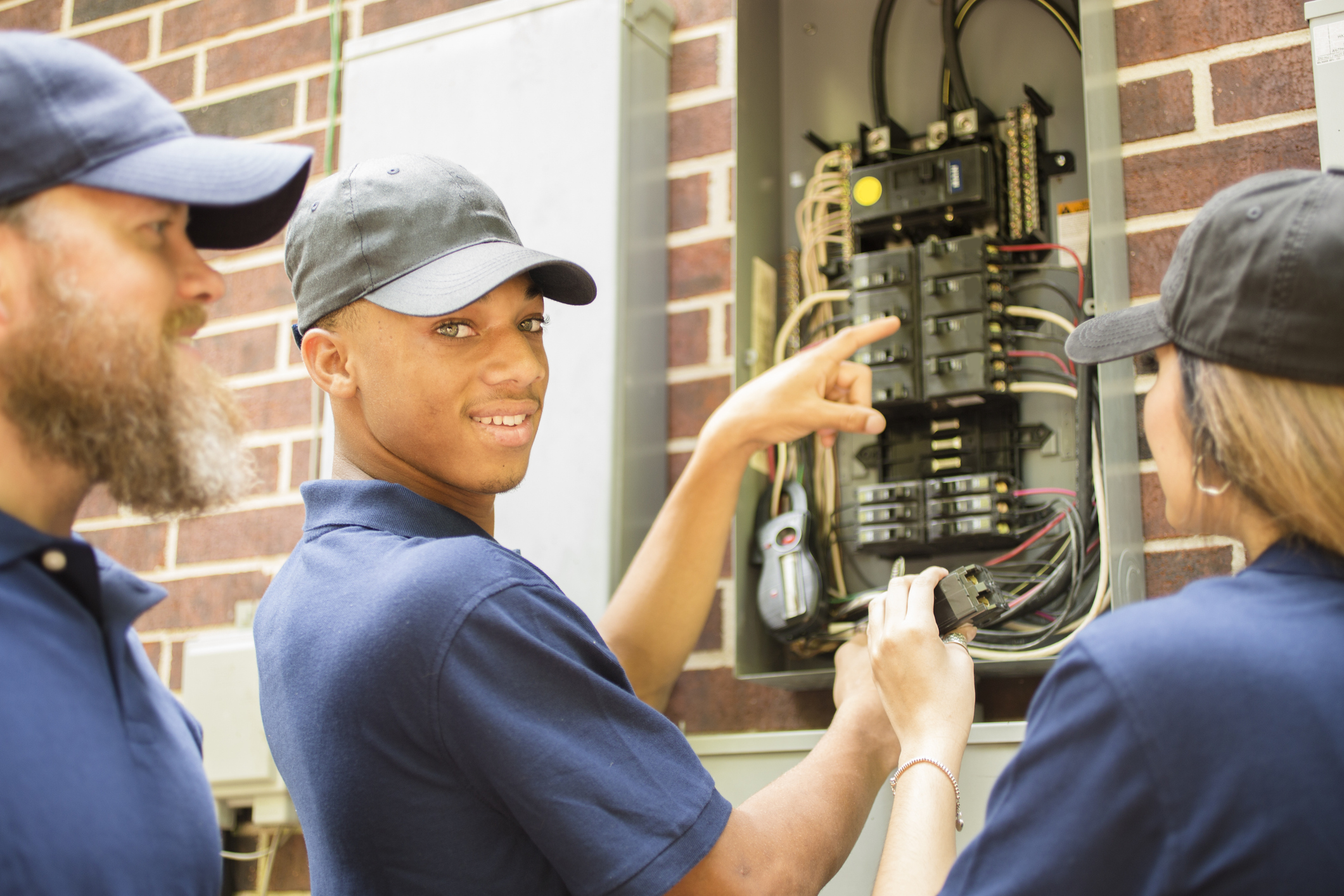 Two men and one woman electricians working on a residential breaker box.  They use tools to assess the repair and wear matching blue uniforms.  The multi-ethnic group is discussing next steps in the job repair.  Electric meter to side.